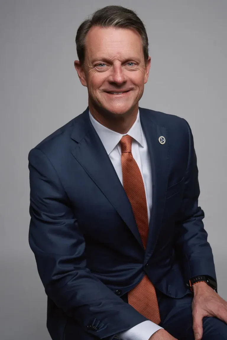 A smiling man in a blue suit, white shirt, and orange tie sits against a plain gray background. He has short brown hair and wears a round pin on his lapel.