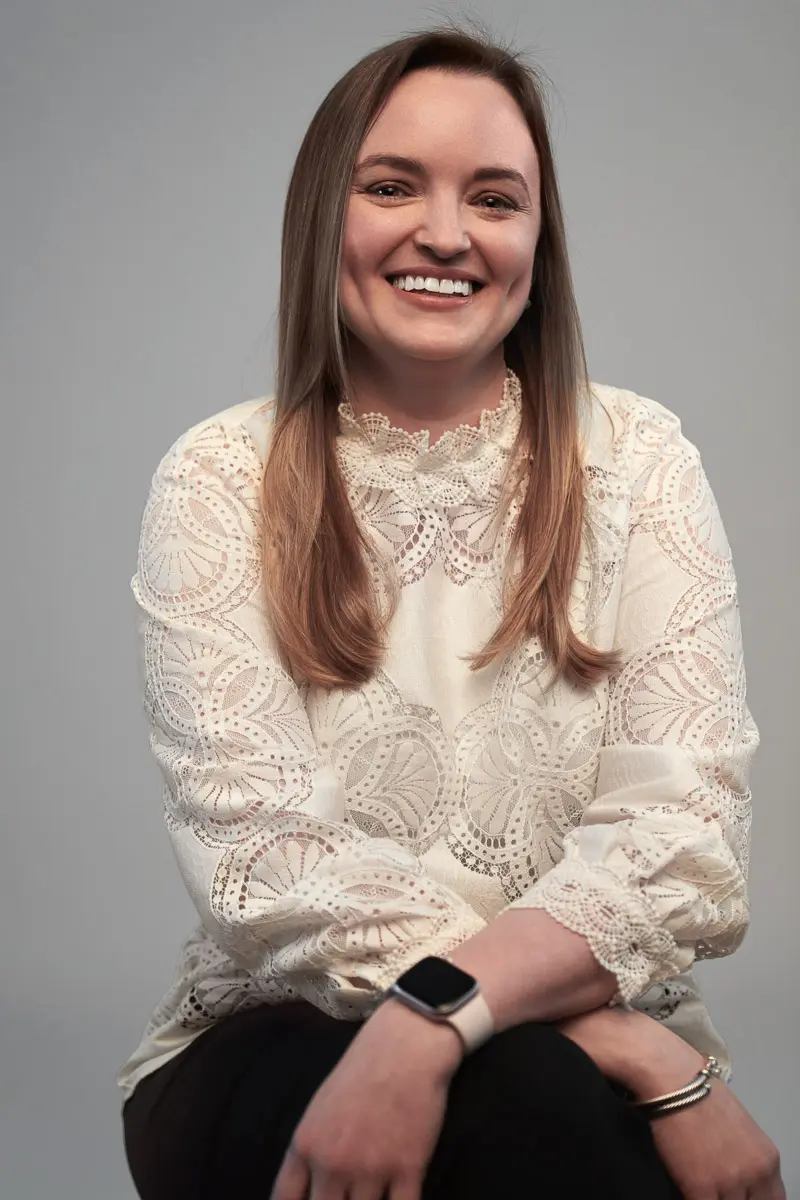 A woman with straight, light brown hair sits smiling, wearing a white lace blouse and a smartwatch on her wrist, with her arms resting casually on her lap against a plain gray background.