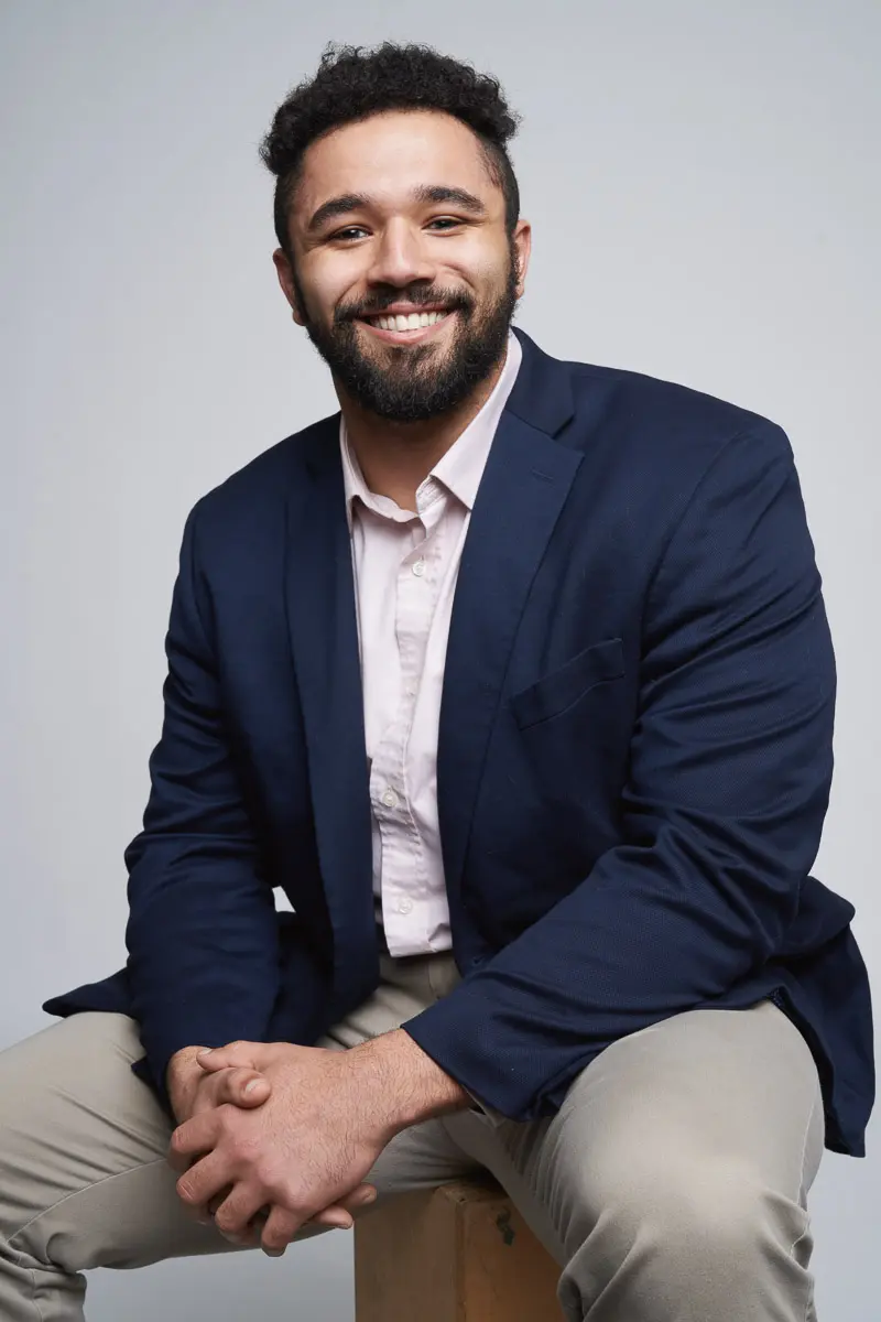 A man with short curly hair and a beard, wearing a navy blazer, light pink shirt, and beige pants, sits smiling against a plain light gray background.