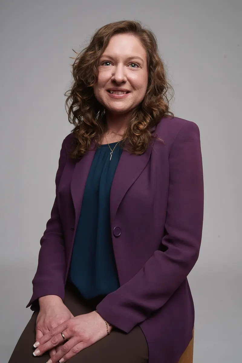 A woman with wavy brown hair wearing a purple blazer over a dark blouse is sitting on a stool against a plain gray background, smiling at the camera.
