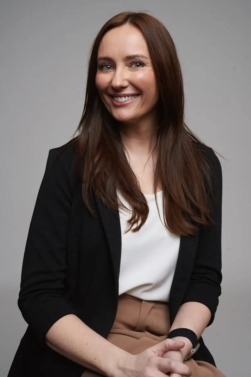 A woman with long brown hair, wearing a black blazer, white blouse, and tan pants, sits and smiles at the camera against a plain gray background.
