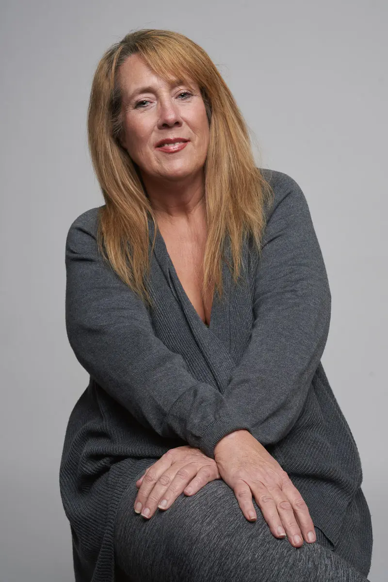 A middle-aged woman with long, light brown hair sits against a plain gray background, wearing a cozy gray sweater and pants. She smiles gently with her hands resting on her knee.