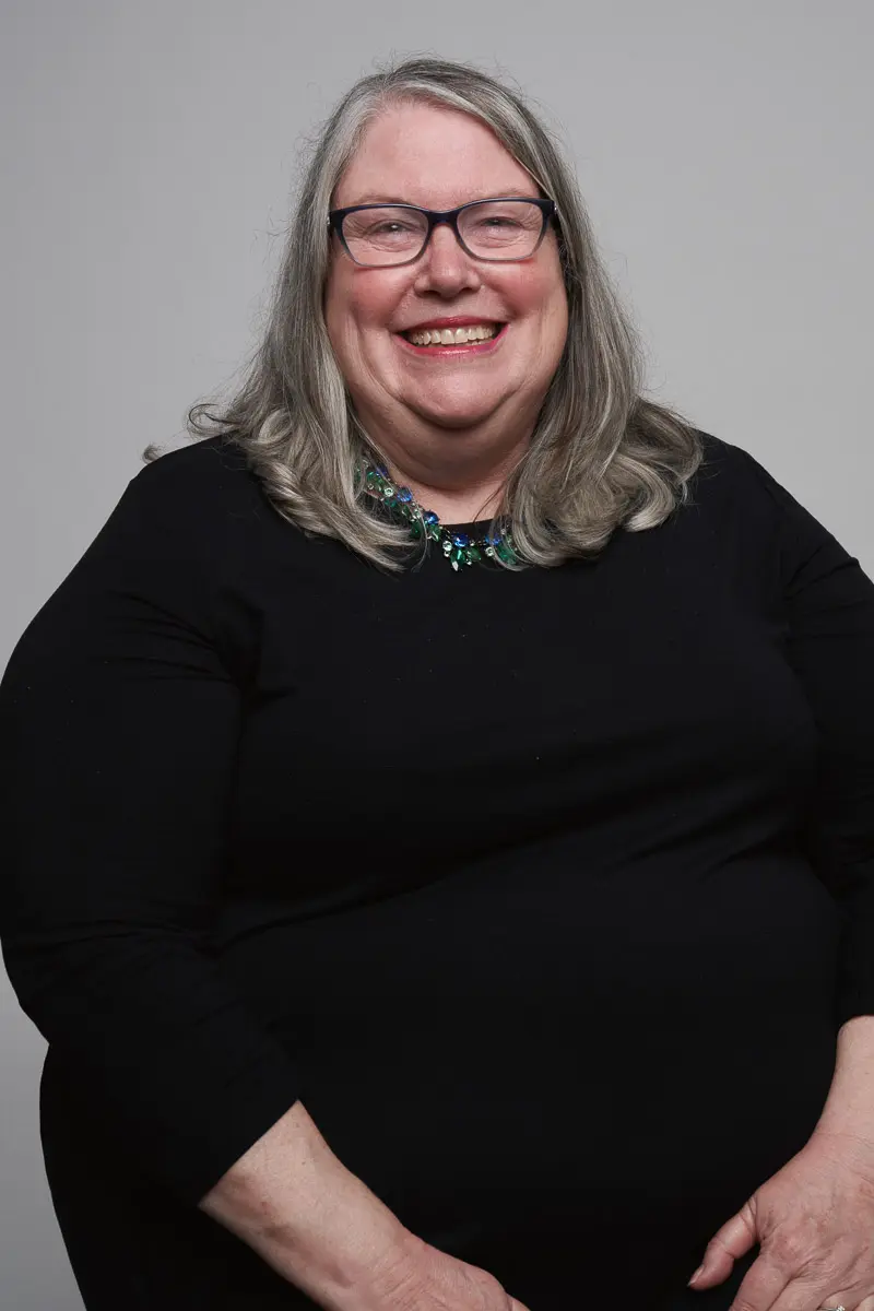 A smiling older woman with long gray hair and glasses wears a black top and a colorful beaded necklace, posing against a plain light gray background.