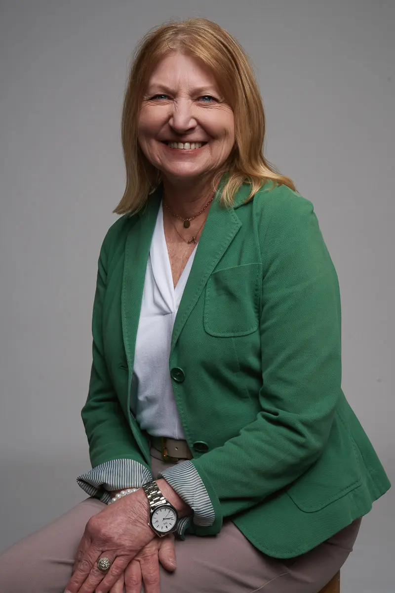 A smiling woman with light brown hair wearing a green blazer, white blouse, and beige pants sits on a stool against a plain gray background. She has a watch on her wrist and hands resting on her lap.