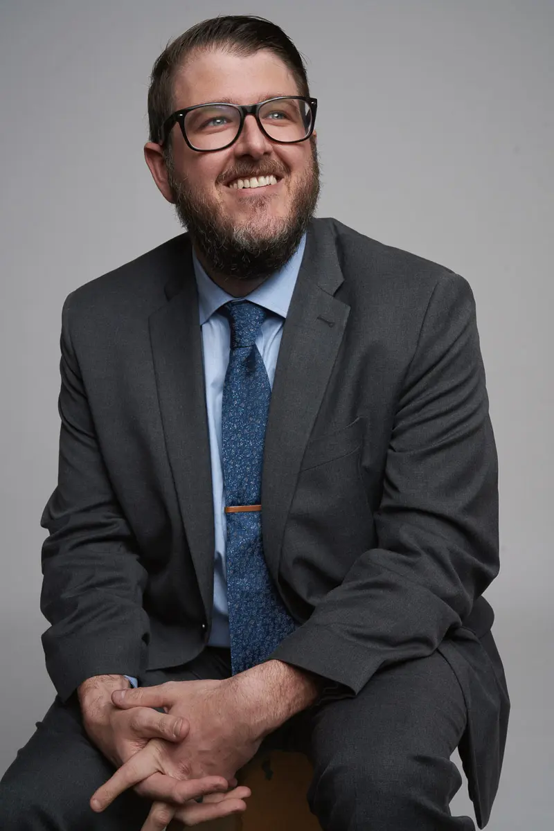 A man with a beard and glasses, wearing a dark suit, light blue shirt, and patterned blue tie, sits on a stool smiling and looking slightly upward against a plain gray background.