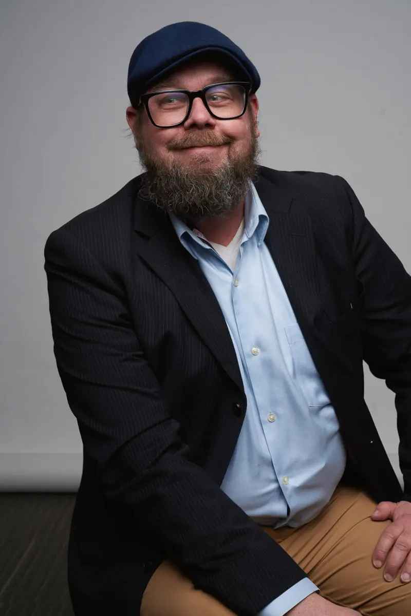 A bearded man wearing a blue flat cap, black glasses, a dark blazer, light blue shirt, and tan pants sits smiling against a plain gray backdrop.