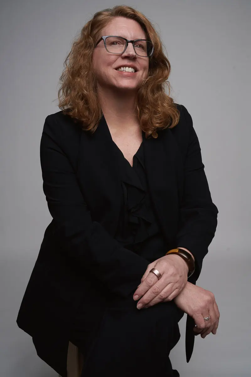 A woman with wavy red hair and glasses smiles while seated, wearing a black suit and resting her hands on her knee against a plain light gray background.