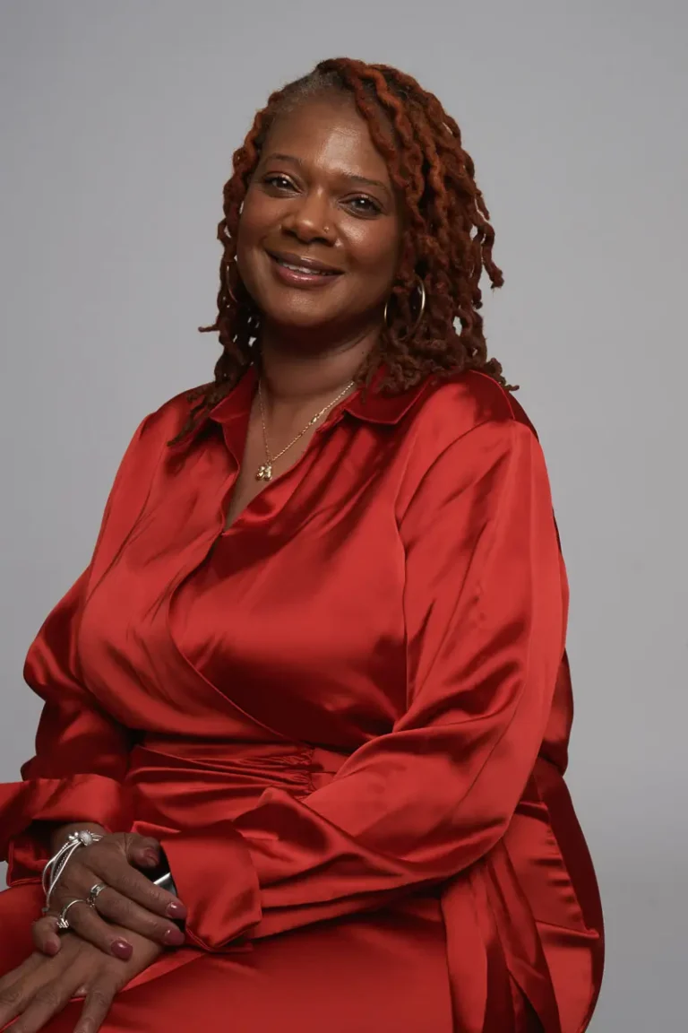 A woman with shoulder-length, twisted reddish-brown hair smiles while sitting. She wears a shiny red wrap dress, gold hoop earrings, necklaces, and bracelets, with her hands resting on her lap against a plain gray background.