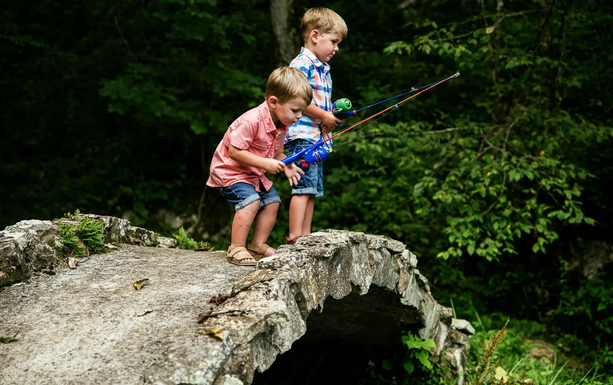 Two young boys in casual clothes stand on a stone bridge, holding fishing rods and looking down, surrounded by lush green trees.