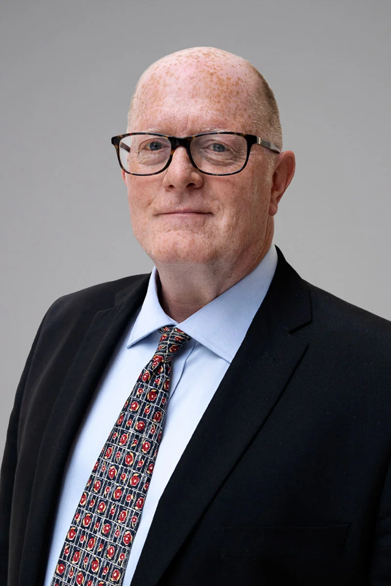 A middle-aged man with light skin, short red hair, and freckles, wearing glasses, a dark suit jacket, a light blue shirt, and a patterned tie, poses against a plain gray background.