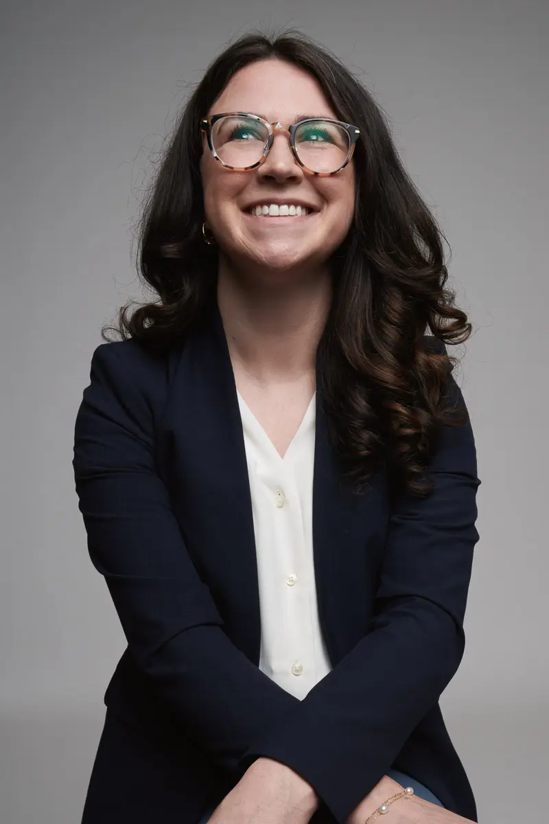 A woman with long, dark, wavy hair and glasses smiles while wearing a dark blazer and white blouse, posing with arms crossed in front of a plain gray background.