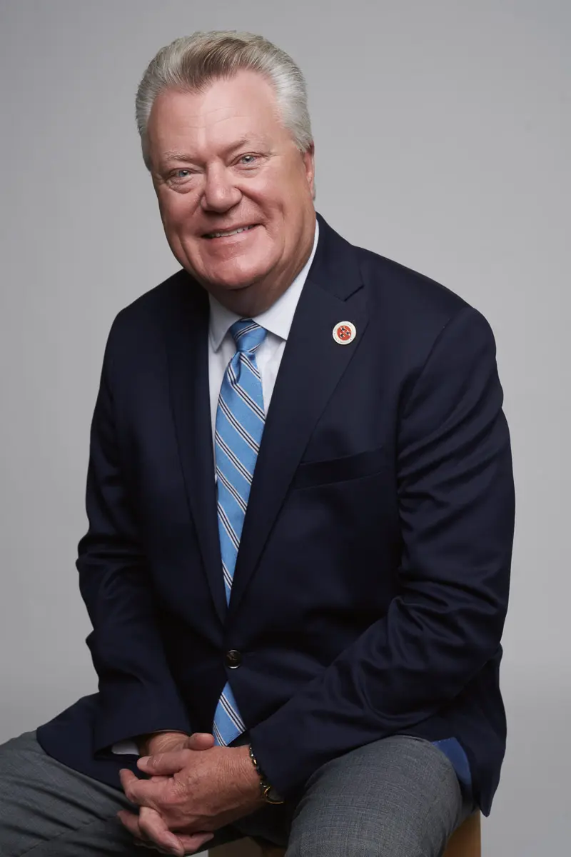 A smiling older man with short gray hair wearing a navy suit, light blue striped tie, and a white dress shirt, sits on a stool against a plain gray background.