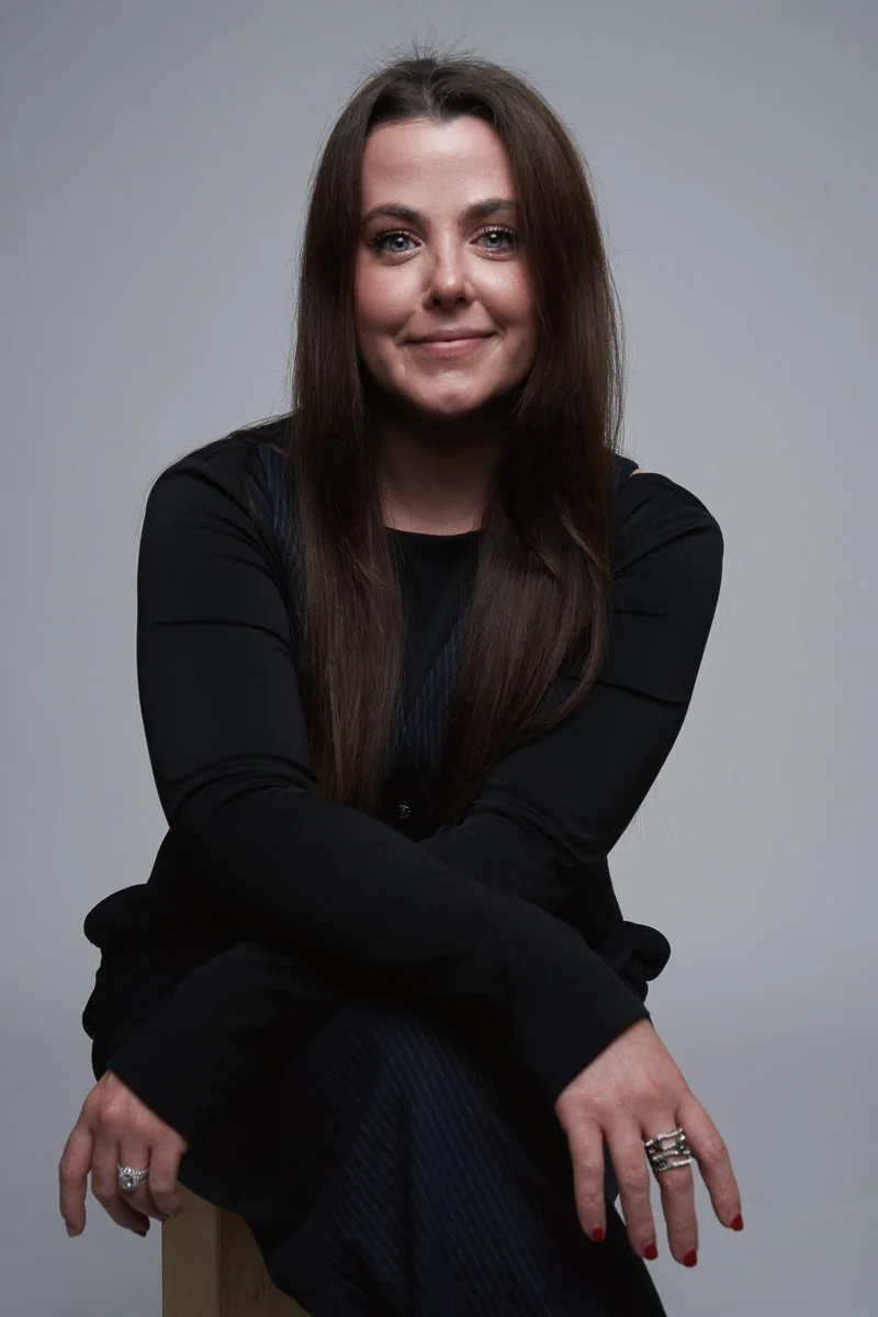A woman with long brown hair and wearing a black outfit sits on a stool with her arms resting on her knees, smiling softly at the camera against a plain gray background.
