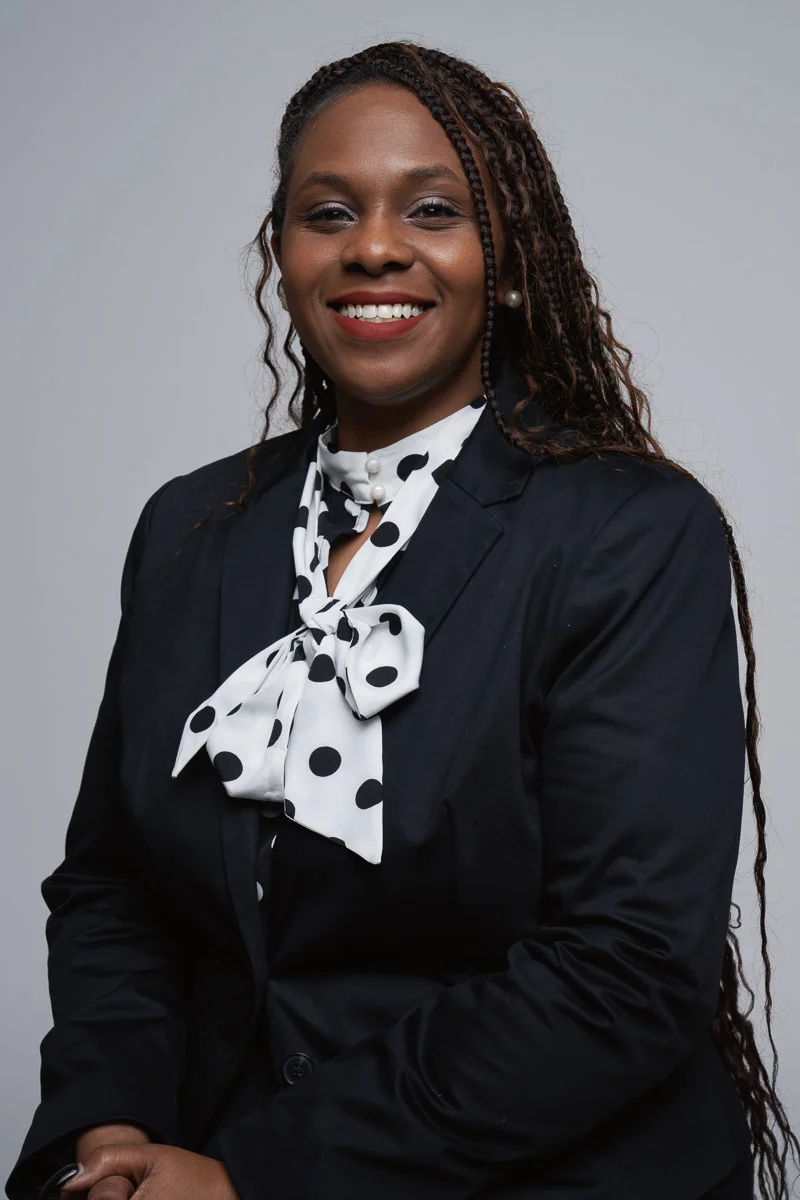 A woman with long braided hair wearing a black blazer and a white blouse with a large black polka dot bow, smiling in front of a plain gray background.