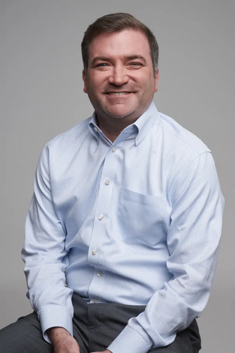 A man with short brown hair smiles while sitting, wearing a light blue button-up shirt and dark pants, against a plain gray background.