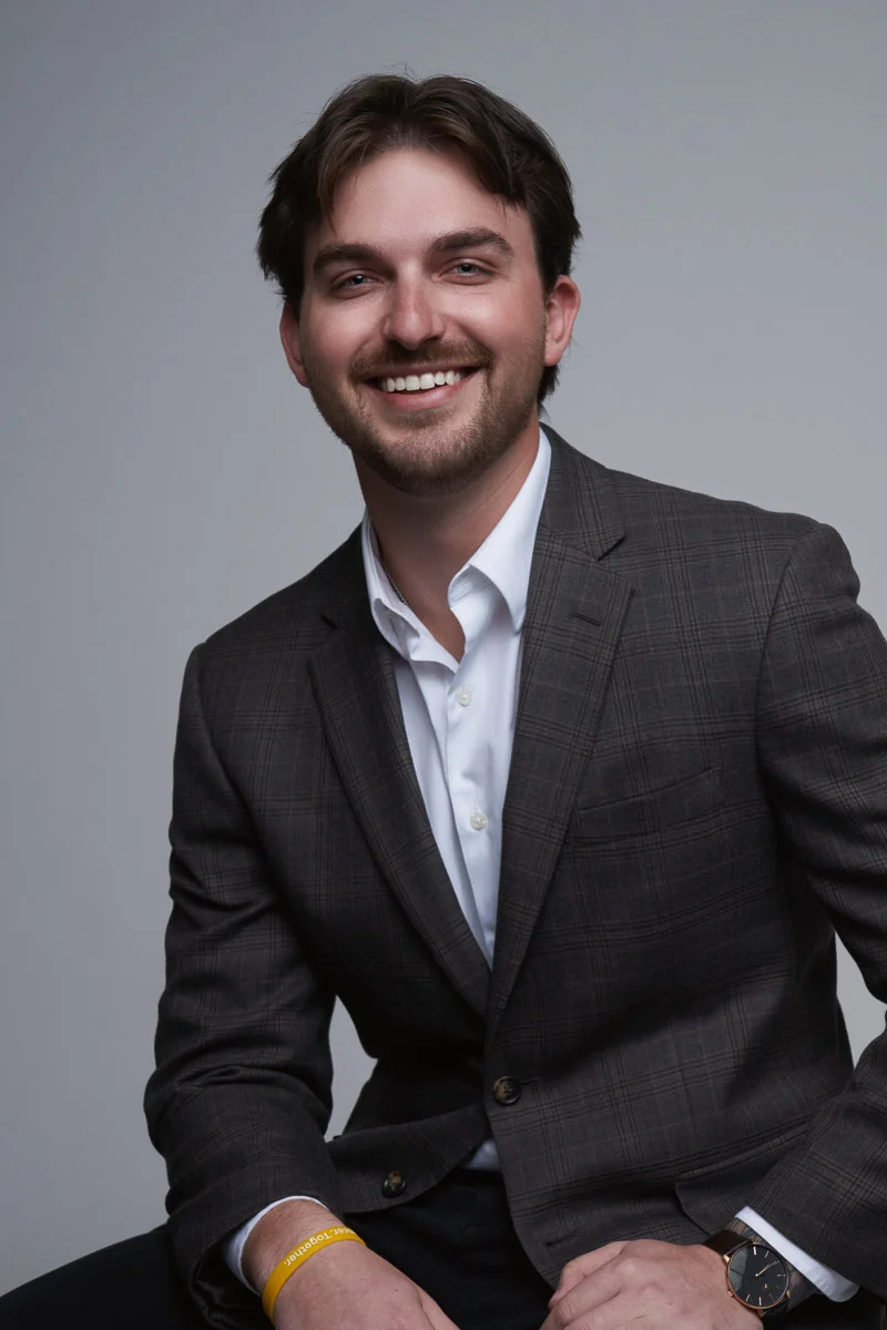 A young man with brown hair and a beard, wearing a gray plaid suit jacket, white shirt, and yellow wristband, sits and smiles against a plain gray background.