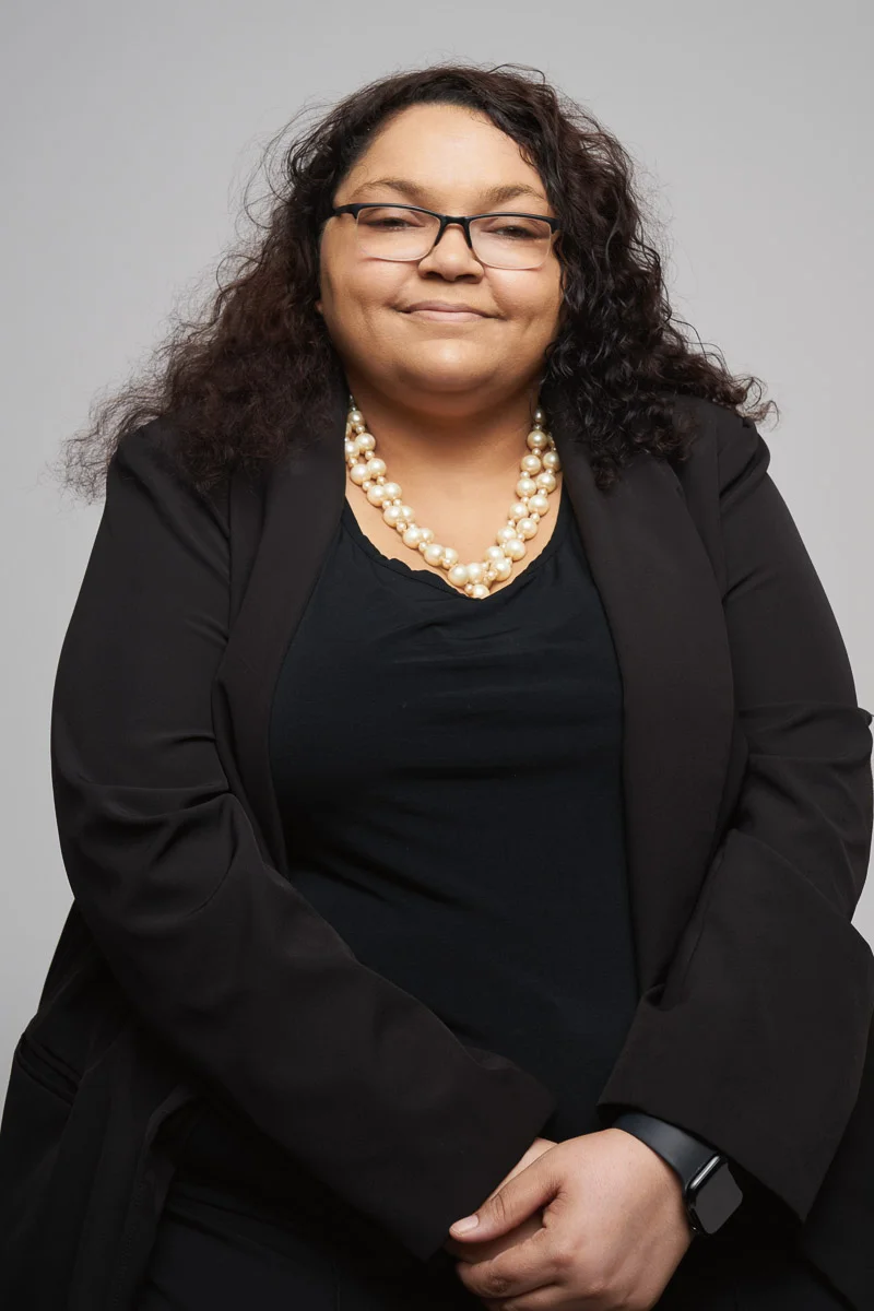 A woman with dark, curly hair wearing glasses, a black blazer over a black top, a pearl necklace, and a smart watch smiles softly while standing with her hands clasped in front of her against a plain gray background.