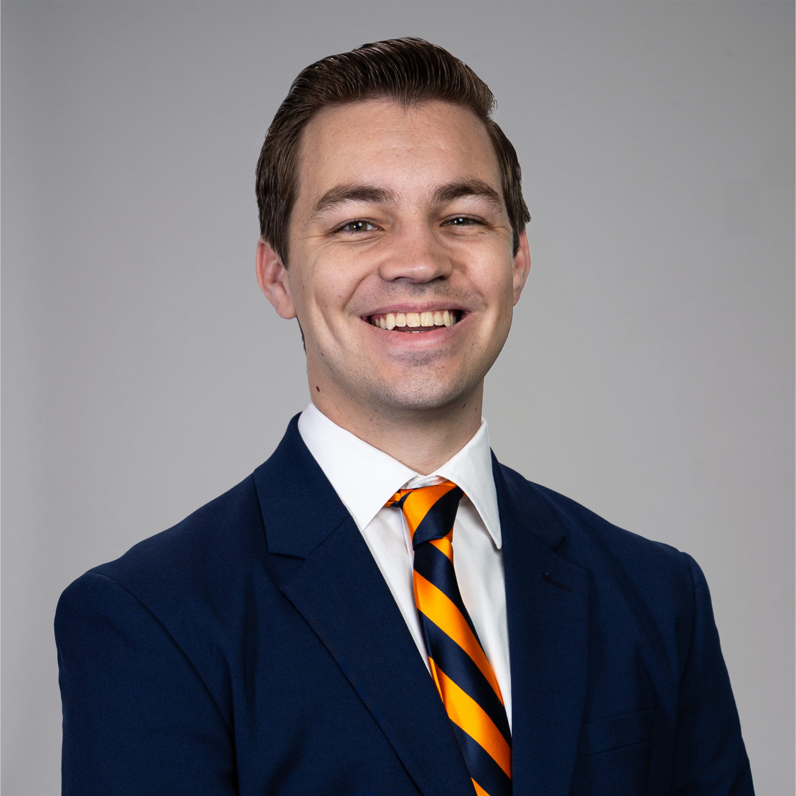A young man with short brown hair smiles while wearing a dark blue suit, white shirt, and a striped orange and navy tie, posing in front of a plain light gray background.