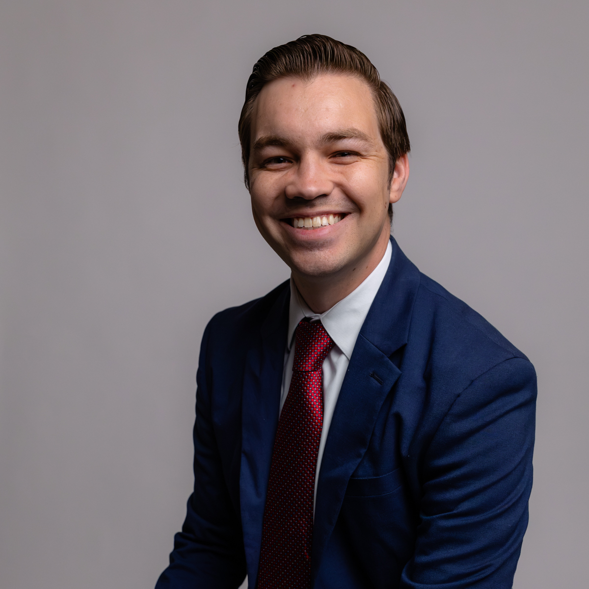 A man in a blue suit and red tie smiles at the camera while sitting against a plain, light gray background.