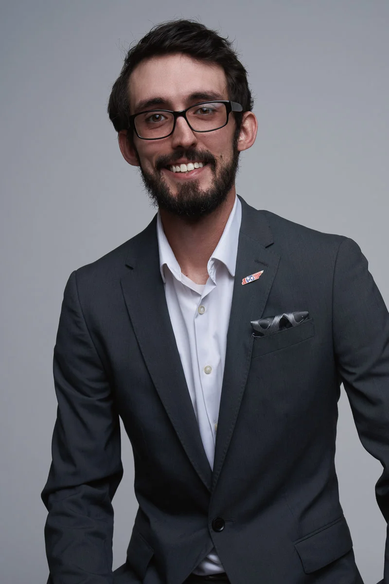 A man with short dark hair, a beard, and glasses smiles while wearing a dark suit, white shirt, and a pocket square, posing in front of a plain gray background.