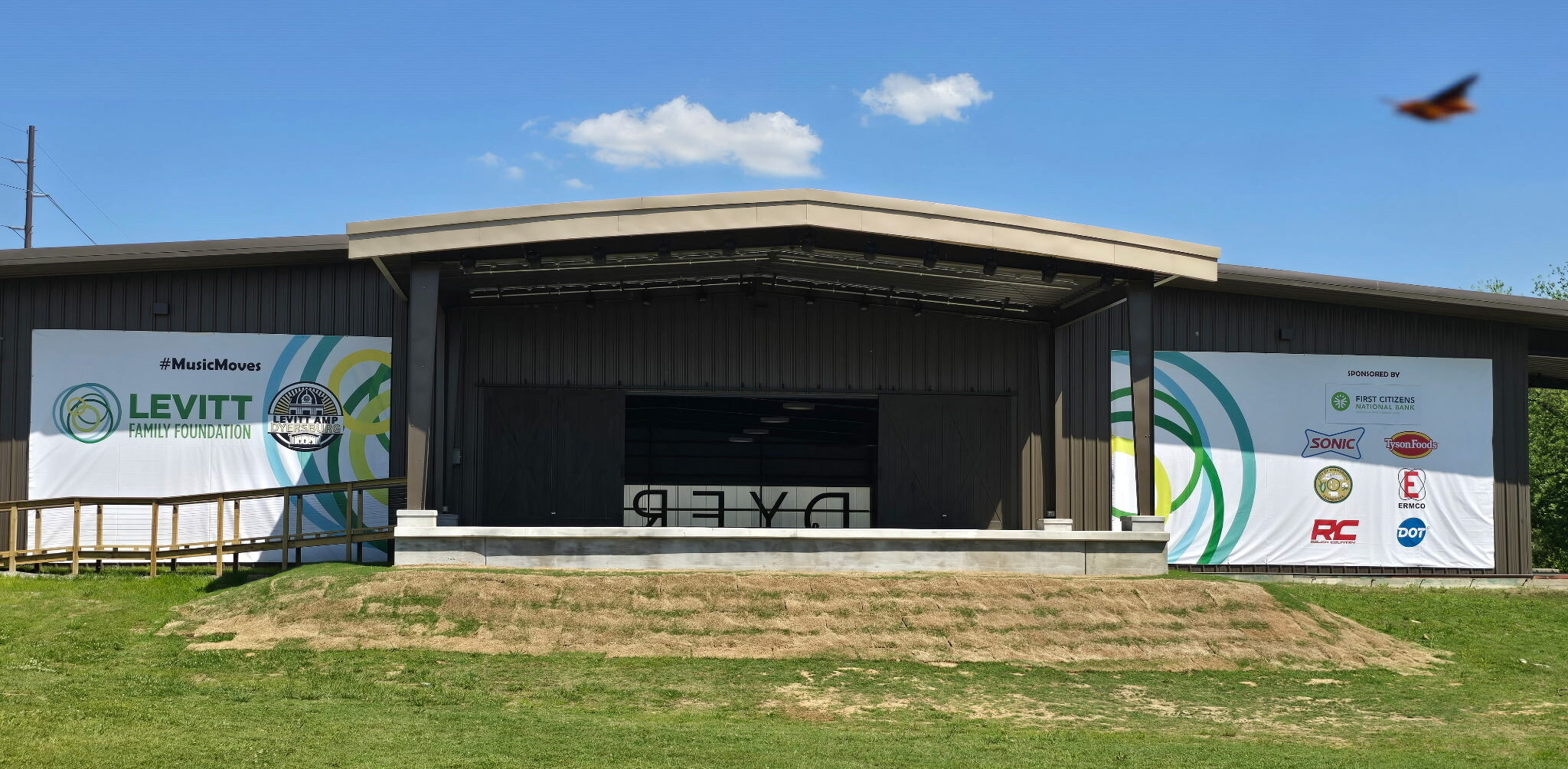 An outdoor stage with a brown roof and banners displaying various logos stands on a grassy area under a blue sky. "DYER" is visible on the stage floor, and a bird is flying in the upper right corner of the image.