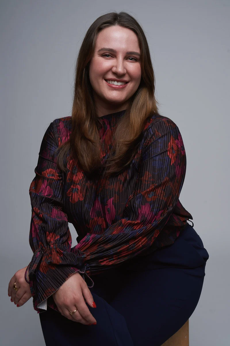 A woman with long brown hair smiles while seated on a stool. She wears a dark, patterned blouse with red and purple accents and navy pants, posing with her hands resting on her knee against a plain gray background.