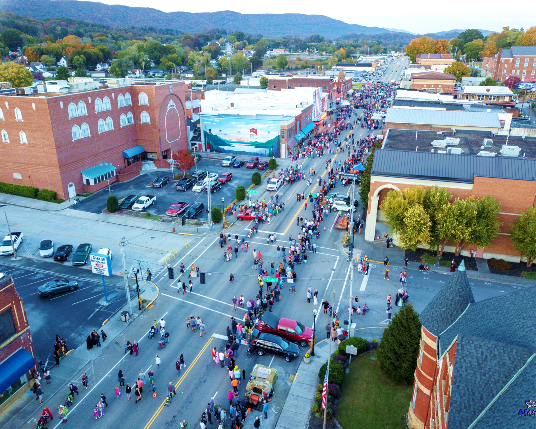 Aerial view of a busy downtown street with crowds of people, many in costumes, walking along both sidewalks and crossing the road, surrounded by brick buildings and parked cars on an autumn day.