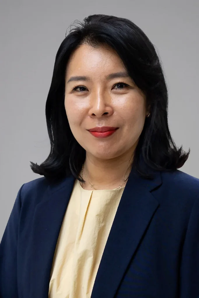 A woman with medium-length black hair, wearing a navy blazer over a light yellow top, smiles softly at the camera against a plain gray background.