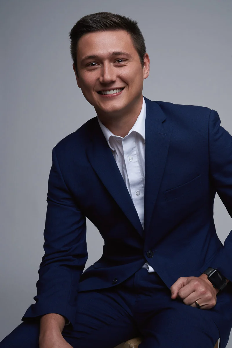 A young man in a navy blue suit and white shirt sits on a stool, smiling at the camera. He has short brown hair and is wearing a smart watch on his left wrist. The background is plain and neutral.