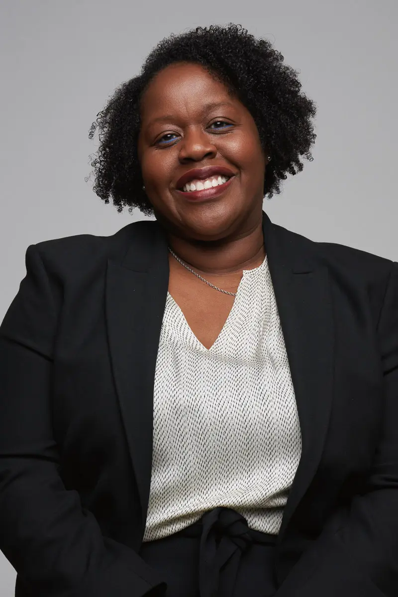 A woman with short curly hair, wearing a black blazer over a light patterned blouse, smiles at the camera against a plain gray background.