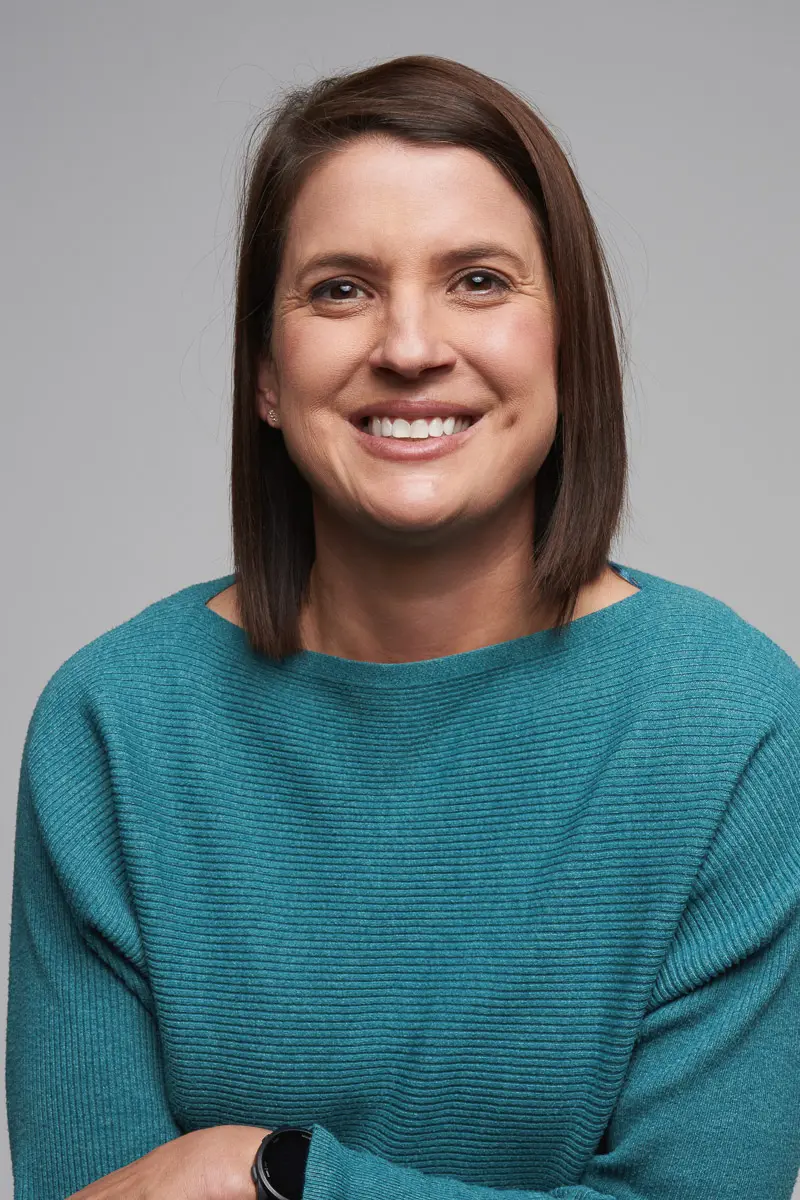 A woman with straight brown hair, wearing a teal sweater and a black watch, smiles at the camera against a plain gray background.