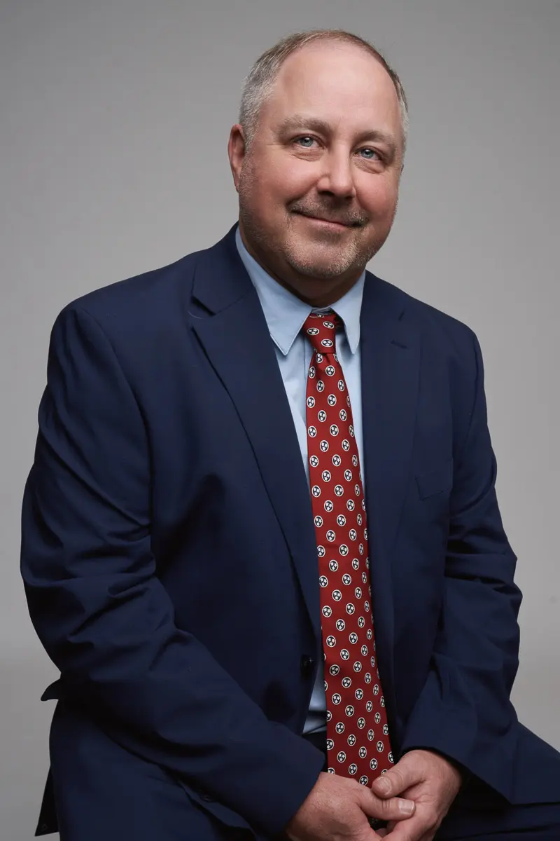 A middle-aged man with short hair, wearing a dark blue suit, light blue shirt, and a red patterned tie, sits and smiles slightly in front of a plain gray background.