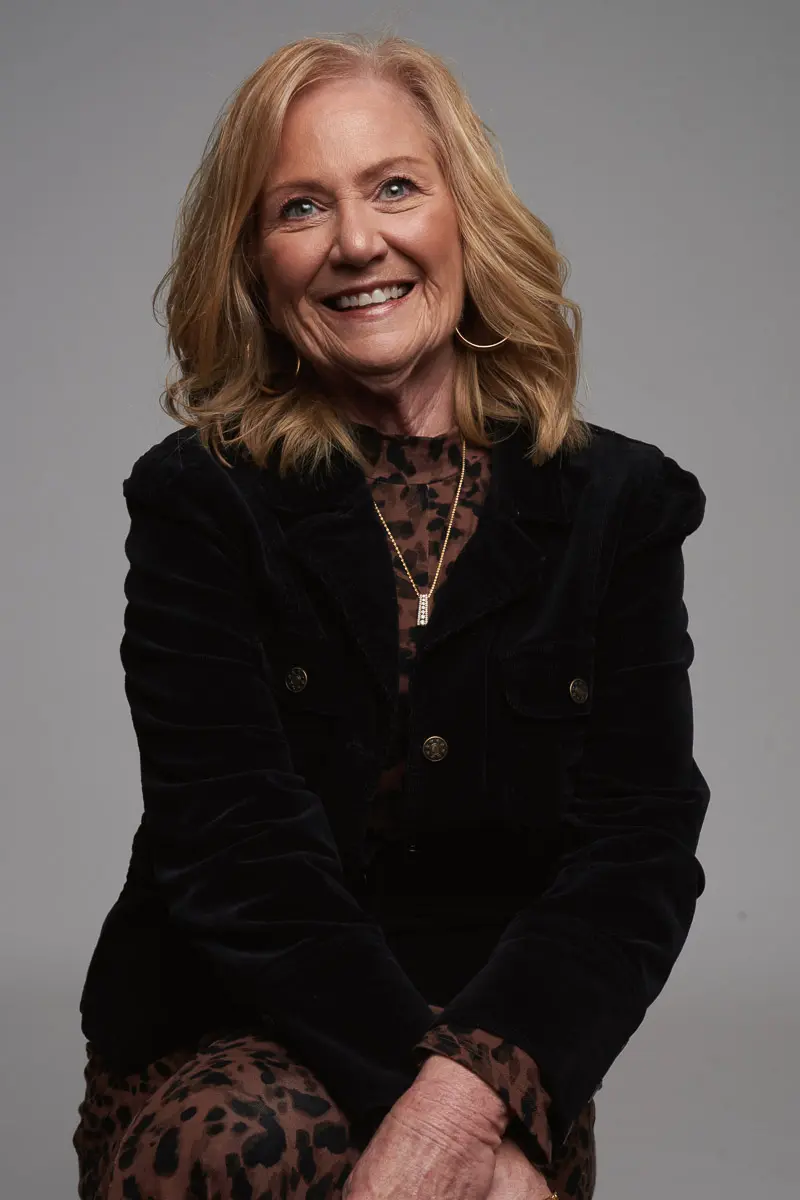 A smiling older woman with shoulder-length blonde hair wears a black jacket over a leopard-print top, sitting against a plain gray background.