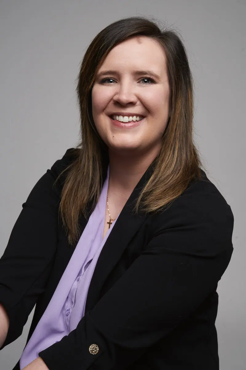 A woman with straight brown hair, wearing a black blazer over a light purple blouse and a cross necklace, smiles at the camera against a plain gray background.