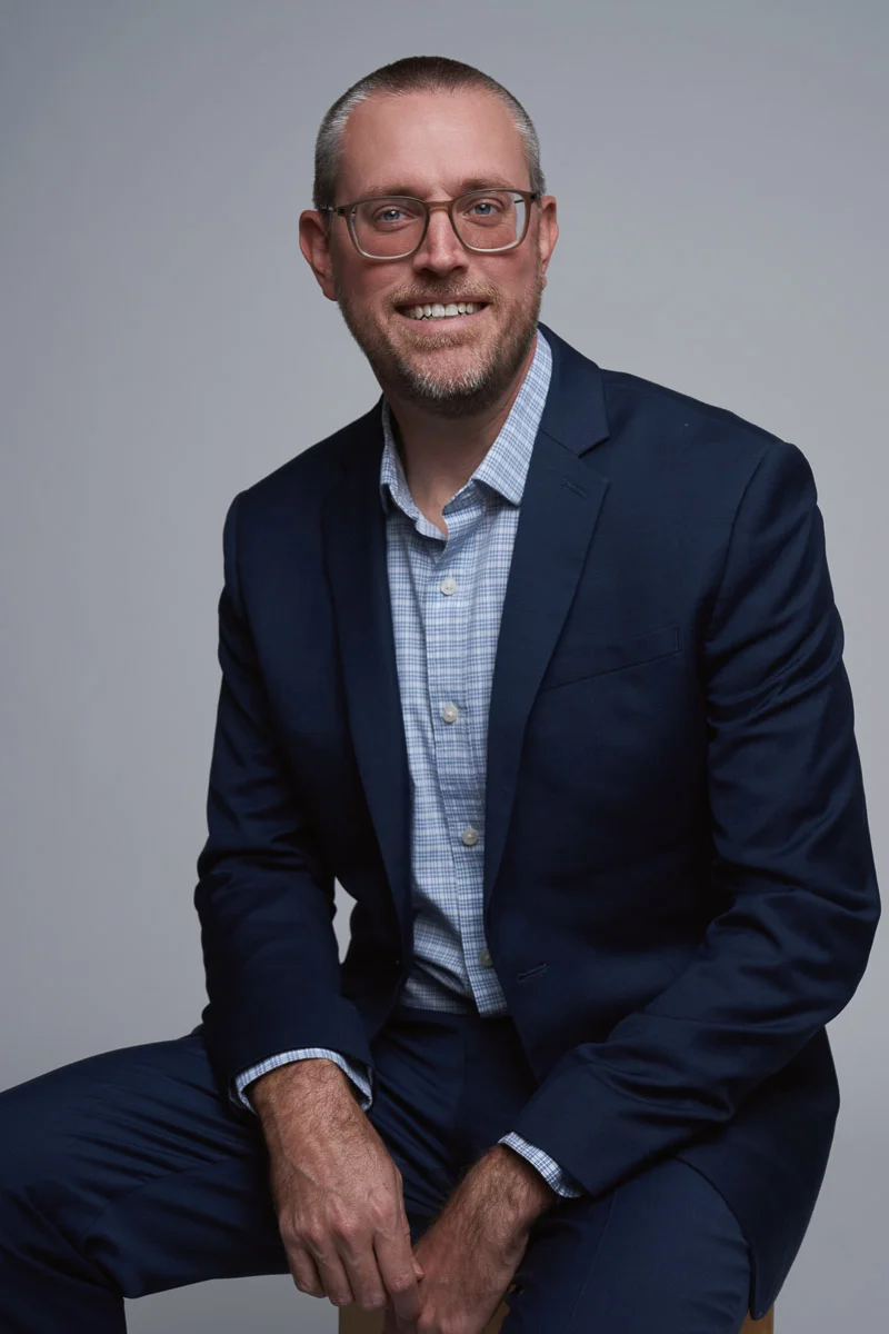 A man with short hair and glasses wearing a navy blue suit and a light blue checked shirt smiles while seated against a plain gray background.