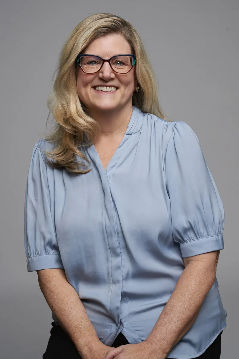 A woman with long blonde hair, wearing glasses and a light blue short-sleeved blouse, smiles while sitting against a plain gray background.