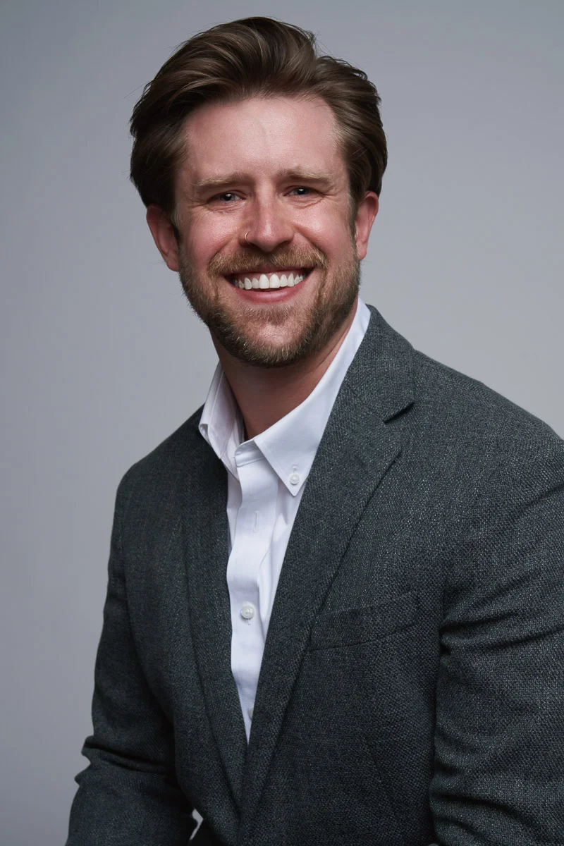 A man with light brown hair and a beard, wearing a white shirt and gray blazer, smiles while posing against a plain gray background.