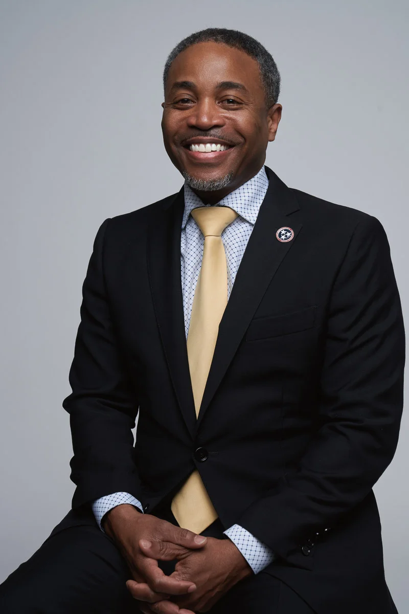 A man in a black suit with a pale yellow tie sits and smiles against a plain light gray background. He wears a dotted dress shirt and a circular pin on his lapel.