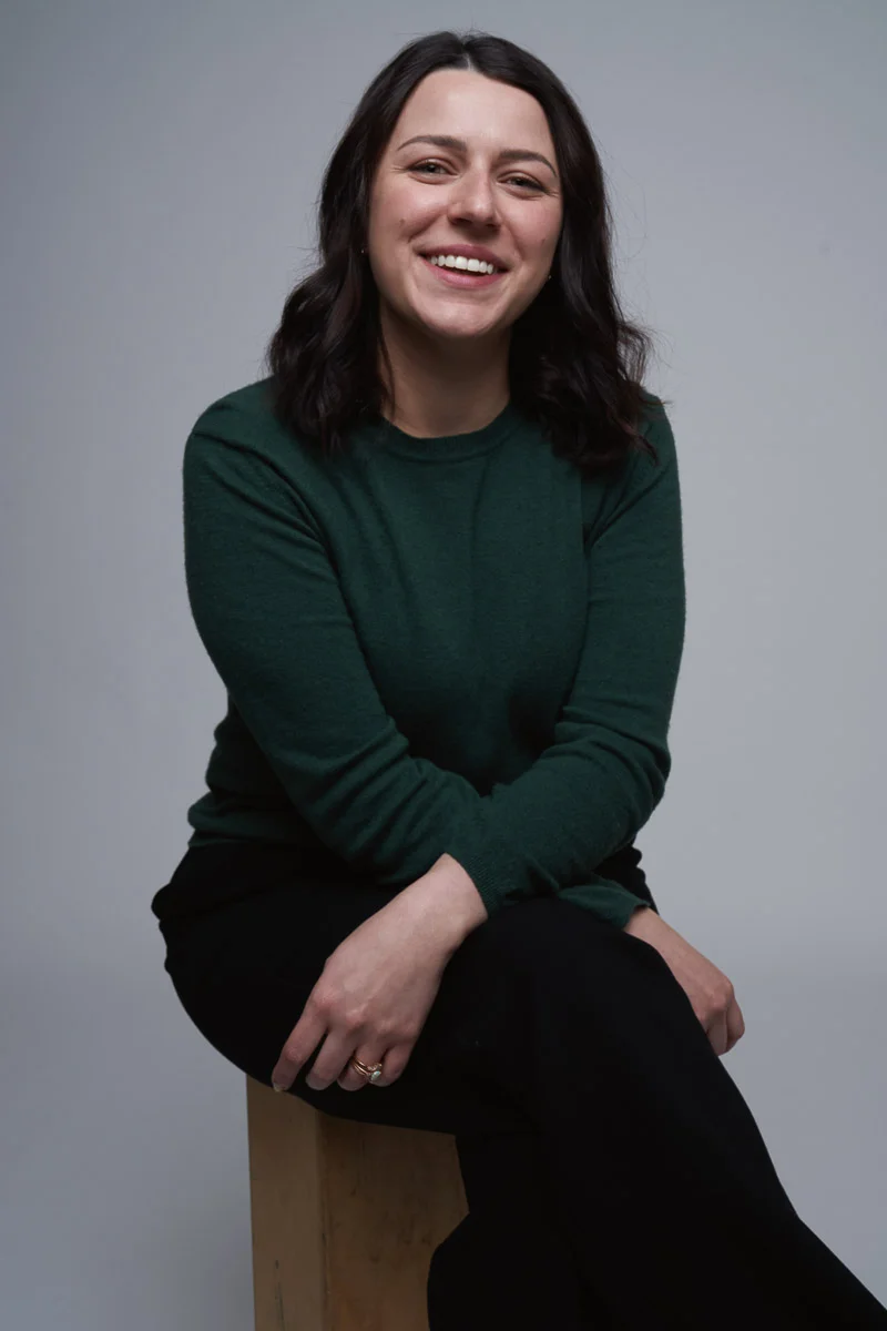 A woman with shoulder-length dark hair, wearing a dark green sweater and black pants, sits on a wooden stool against a plain light background, smiling warmly with her hands resting on her knee.