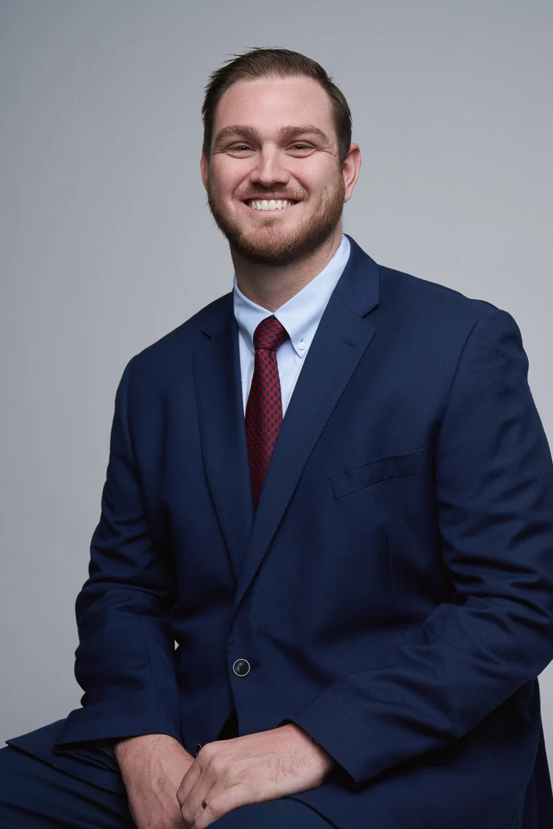 A man with short hair and a beard, wearing a navy blue suit, white shirt, and red tie, sits and smiles at the camera against a plain light gray background.