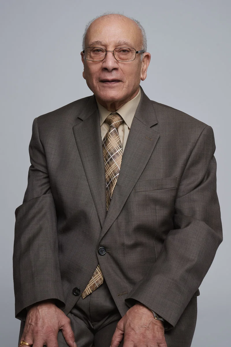 An older man wearing glasses, a brown suit, and a patterned tie poses for a formal portrait against a plain light gray background.