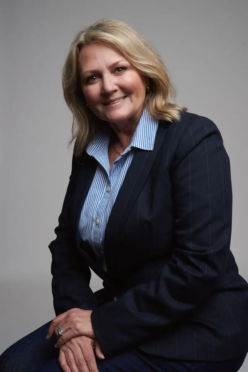 A woman with blonde hair wearing a navy pinstripe blazer and a blue striped shirt sits and smiles at the camera against a plain gray background.
