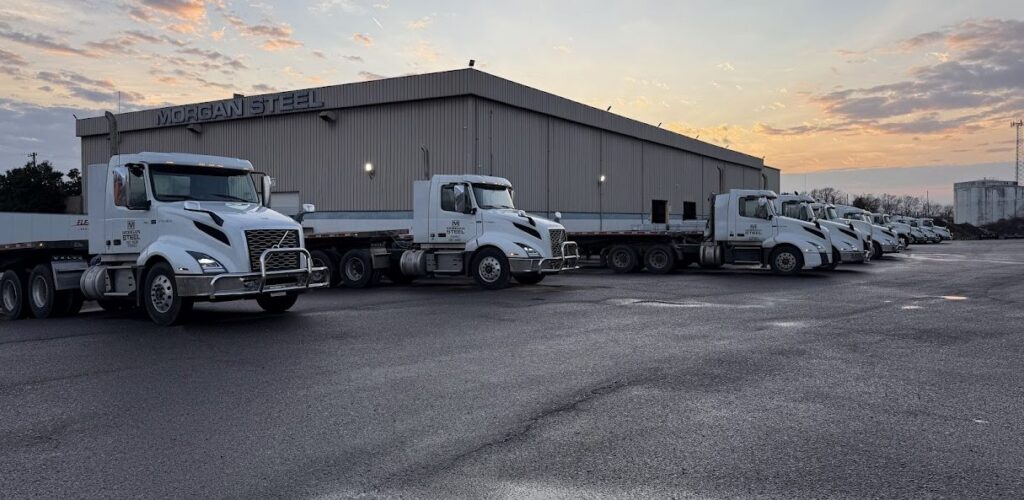 Several white semi-trailer trucks are lined up in front of a large industrial building labeled "Morgan Steel" in the early morning or at sunset, with a cloudy sky overhead.