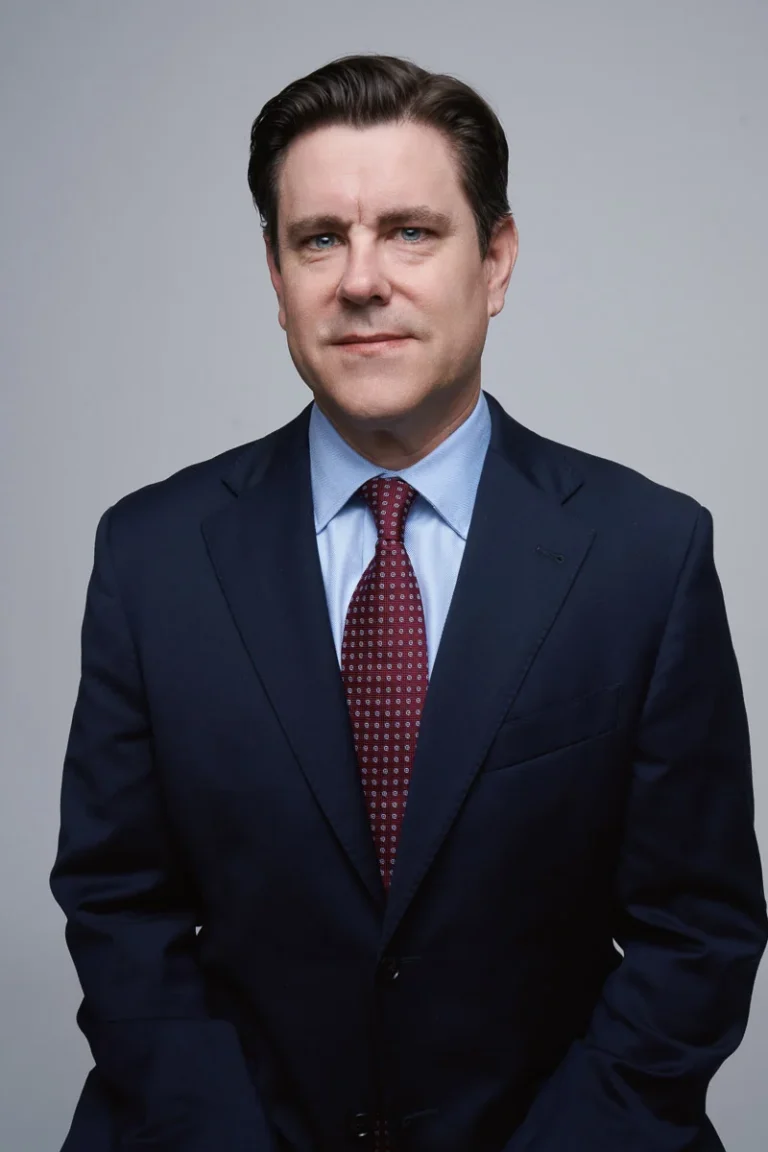 A man with brown hair wearing a navy blue suit, light blue dress shirt, and patterned red tie poses against a plain gray background, looking directly at the camera with a neutral expression.