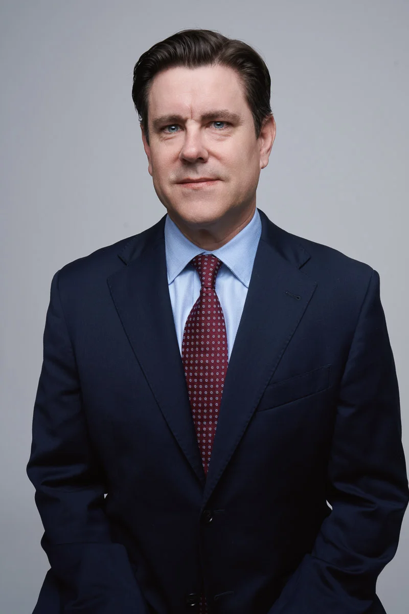 A man with brown hair wearing a navy blue suit, light blue dress shirt, and patterned red tie poses against a plain gray background, looking directly at the camera with a neutral expression.