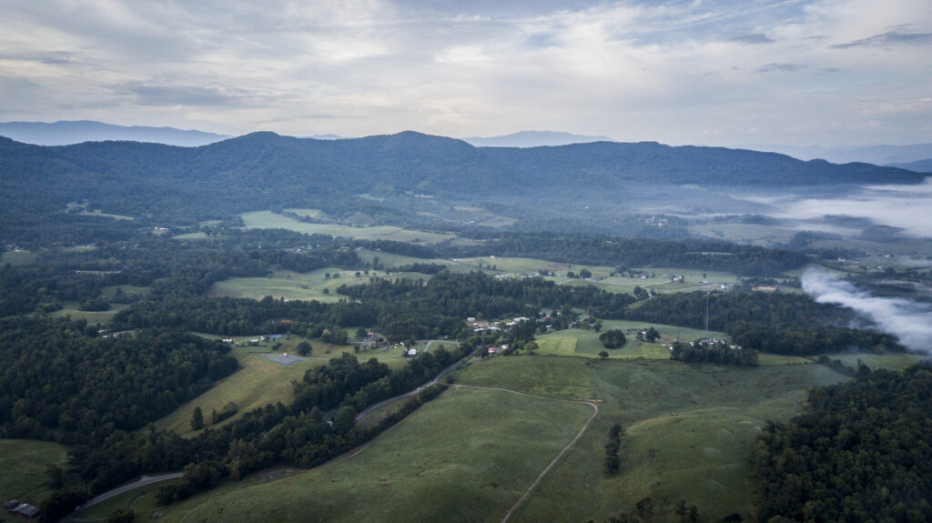 Aerial view of a rural landscape with green fields, scattered houses, dense forests, and rolling hills under a cloudy sky, with mist visible in the distance near the mountains.