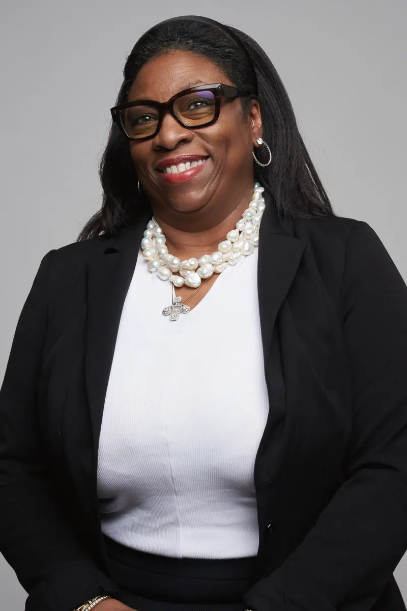 A woman with long dark hair, wearing glasses, a black blazer, a white top, and layered pearl necklaces, smiles in front of a plain gray background.