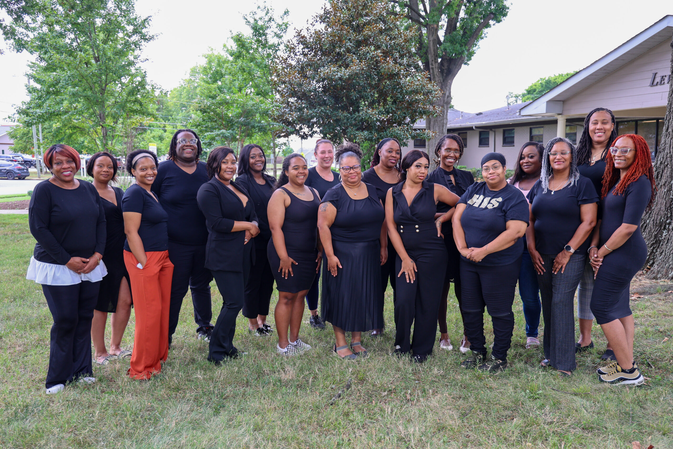 A group of seventeen adults poses together on grass outside a building. Most are wearing black clothing and are smiling at the camera. Trees and houses appear in the background.