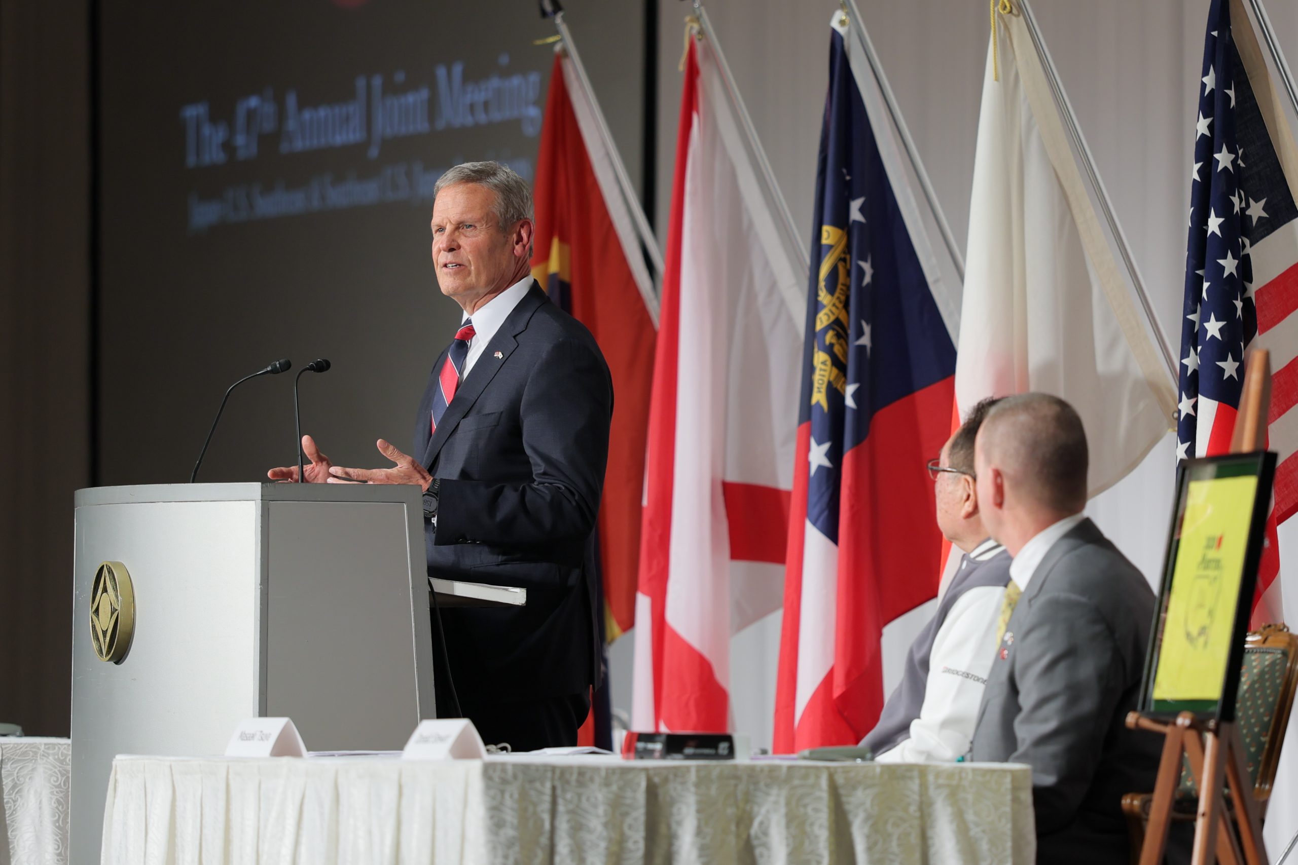 A man in a suit speaks at a podium during a formal event, with several people seated nearby. Multiple national and state flags are displayed in the background on a stage.