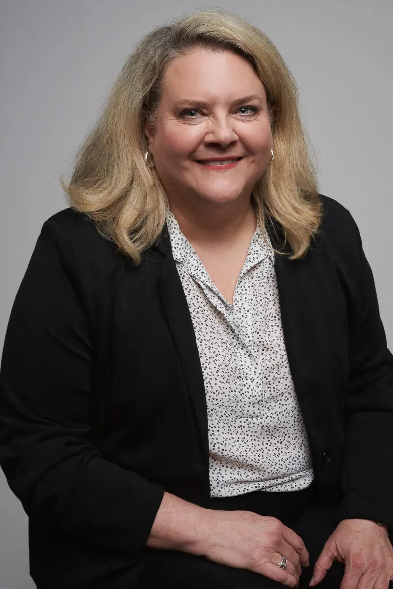 A woman with shoulder-length blonde hair, wearing a black blazer over a white patterned blouse, sits smiling against a plain gray background.